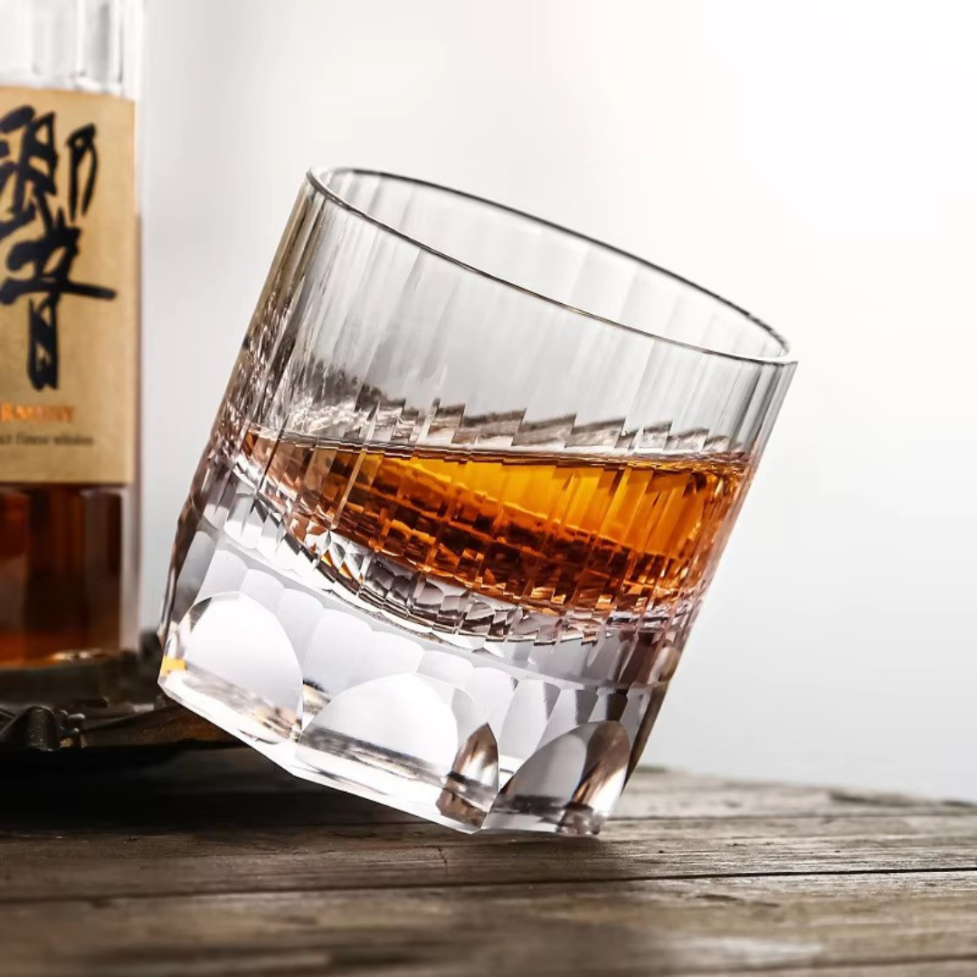 Handcrafted K9 crystal whisky glass with a heavy bottom and intricate kiriko cut design, featuring thumbprint-inspired imprints for a comfortable grip. Slightly tilted with whisky on a rustic wooden bench.