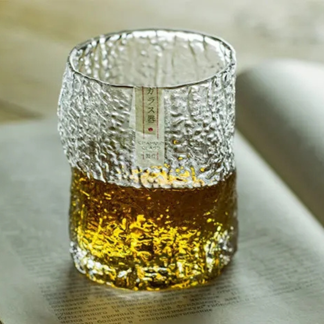 Close-up of a handcrafted Edo whisky glass with a unique crumple pattern, filled with whisky, placed on an open book. The textured crystal surface catches the light, highlighting its artisanal design.